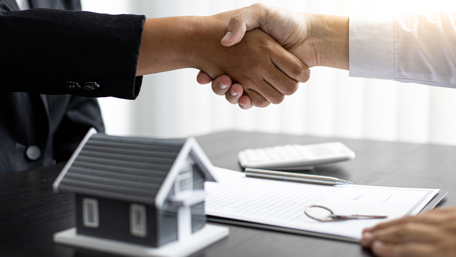 Two hands shaking over a table with a small model house, eyeglasses, a contract, a pen, and a calculator. The scene suggests the closing of a real estate deal or financial agreement, possibly facilitated by experts from an SMSF accountants group or Melbourne accounting firm.