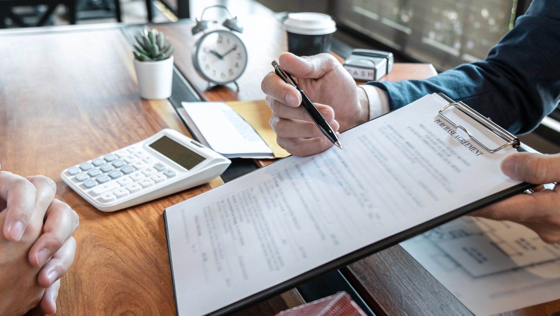 A person is holding and pointing to a clipboard with a document while another person watches. A calculator, clock, coffee cup, and a small plant are on the wooden table in the background. The scene appears to be a meeting or consultation at a Melbourne Accounting Firm.