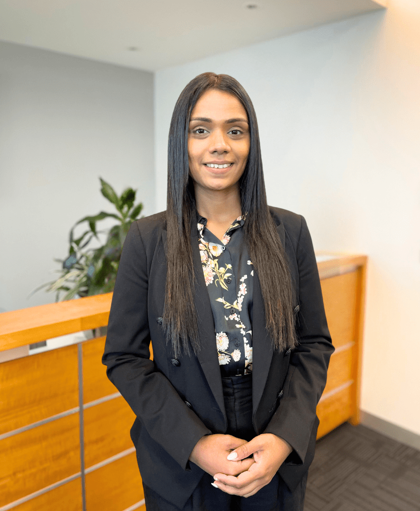 A person with long dark hair is standing in an office space wearing a black blazer and floral blouse. They are smiling, with hands clasped in front. Behind them is a railing and a green plant in the background.