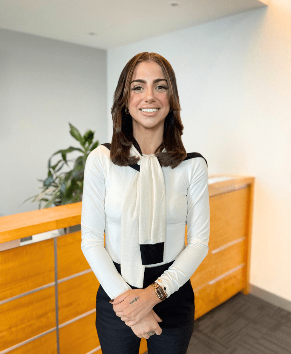 A person with shoulder-length brown hair is smiling and standing in a modern office space. They are wearing a white long-sleeve top with a large scarf and black pants. There are plants and wooden paneling in the background.