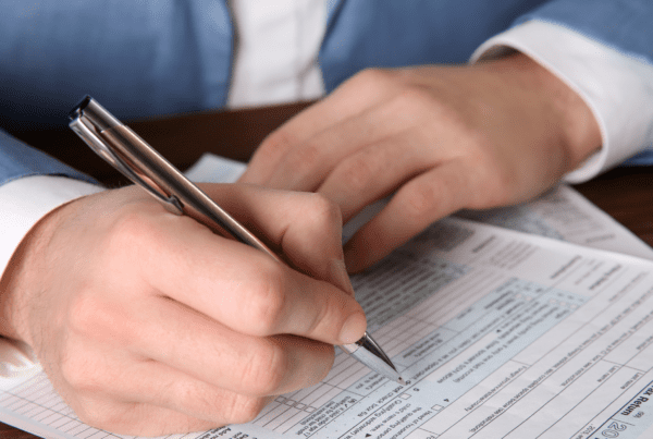 A person in a light blue suit jacket is filling out a form at a wooden desk, possibly seeking tax advice. They hold a pen, and the document has small printed text and boxes. Only the person's hands and part of the arms are visible. This scene is reminiscent of many Melbourne accounting firms specializing in detailed financial matters.