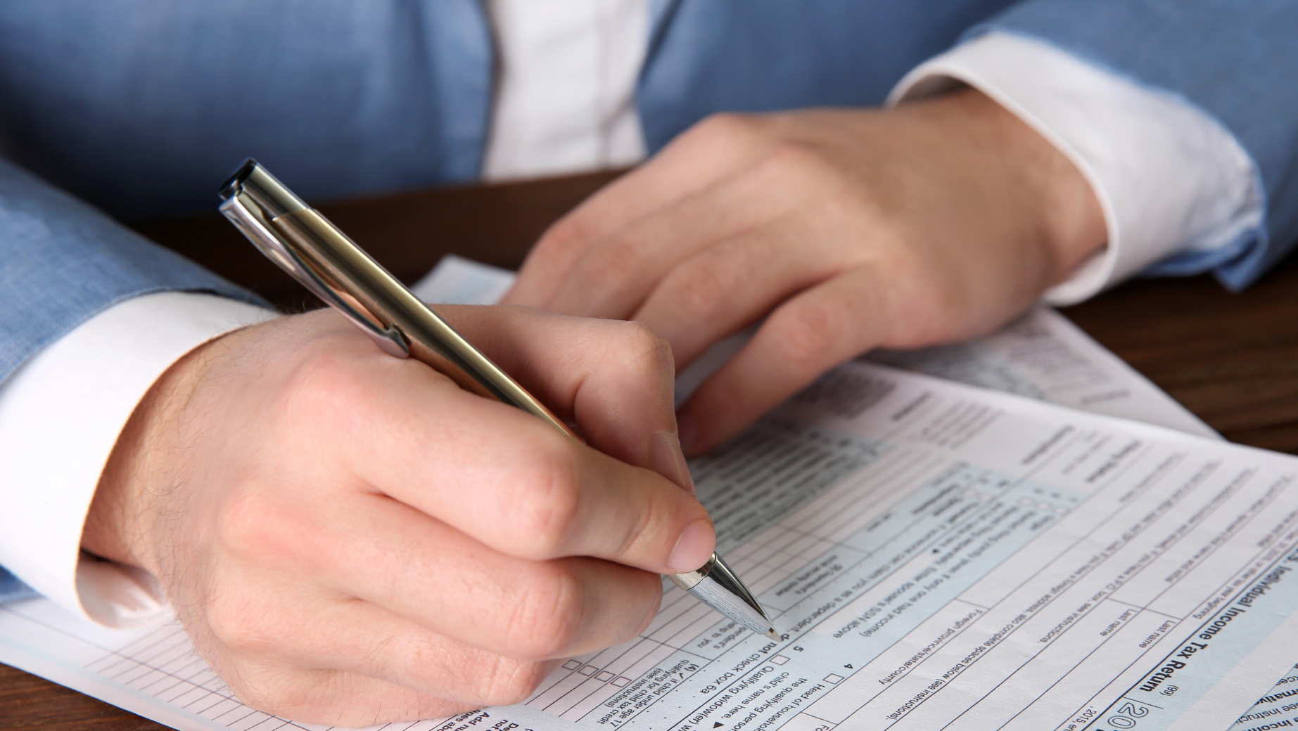 A person in a light blue suit jacket is filling out a form at a wooden desk, possibly seeking tax advice. They hold a pen, and the document has small printed text and boxes. Only the person's hands and part of the arms are visible. This scene is reminiscent of many Melbourne accounting firms specializing in detailed financial matters.