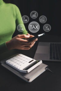 A person in a green sweater uses a smartphone at a desk with a laptop and calculator. Above the phone, digital icons and the word "TAX" are displayed, representing tax-related concepts.