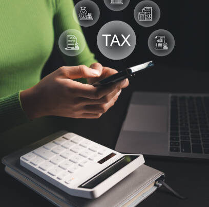 A person in a green sweater uses a smartphone at a desk with a laptop and calculator. Above the phone, digital icons and the word "TAX" are displayed, representing tax-related concepts.