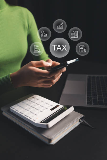 A person in a green sweater uses a smartphone at a desk with a laptop and calculator. Above the phone, digital icons and the word "TAX" are displayed, representing tax-related concepts.