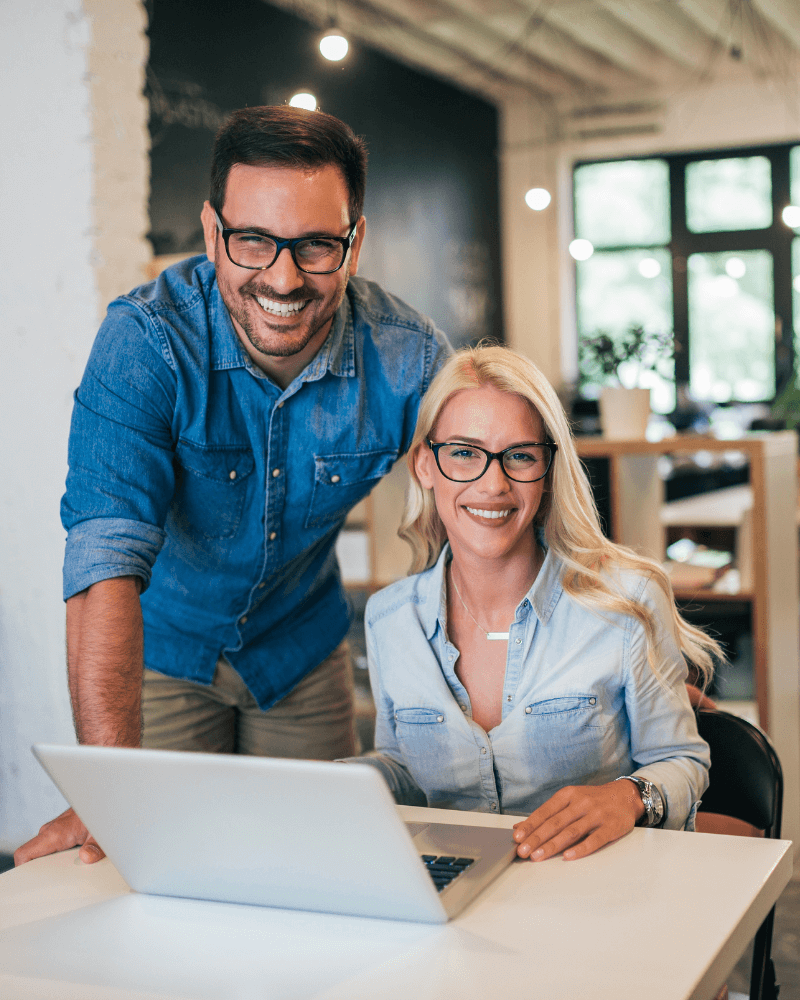 A smiling man and woman wearing glasses and casual denim shirts are working together at a desk with an open laptop. The office setting, part of a Melbourne accounting firm, has a white interior with ambient lighting and large windows. The man stands beside the seated woman, providing tax advice.