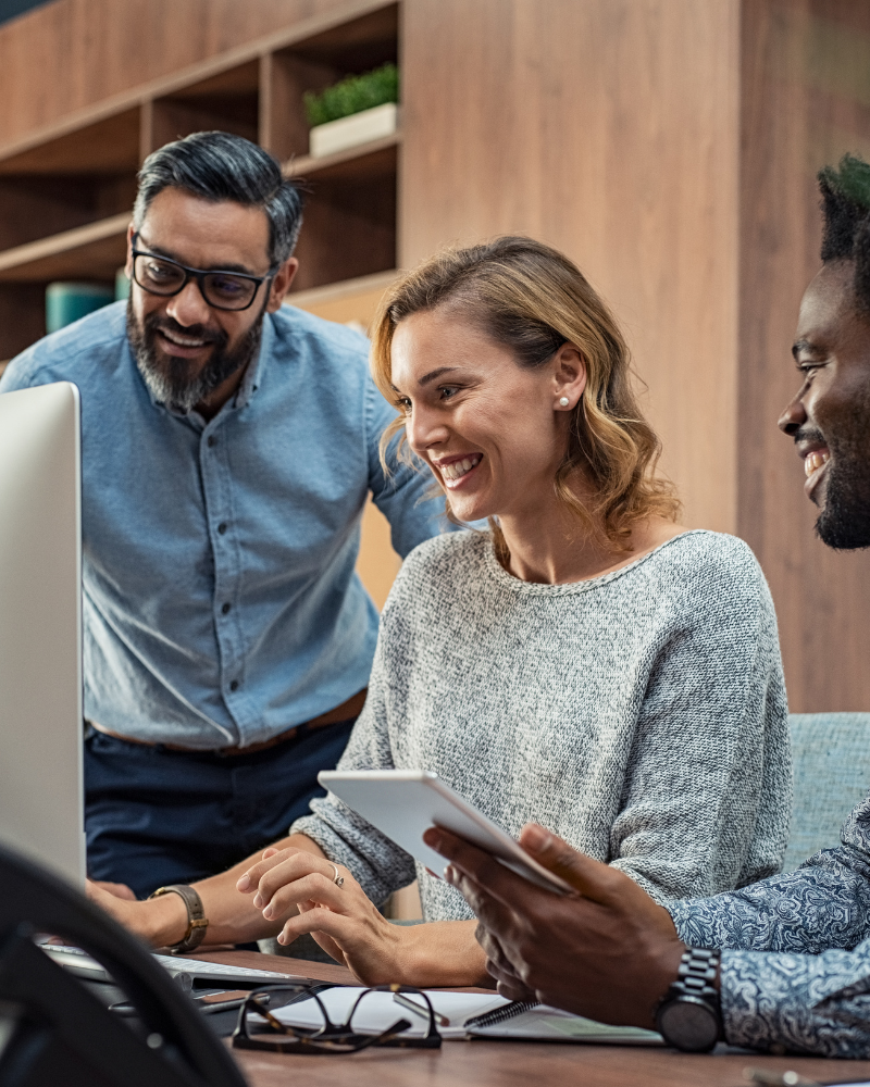 Three colleagues smile while working together at a desk. The woman in the middle types on a keyboard, the man on the left leans over watching, and the man on the right holds a tablet. They appear engaged and happy, collaborating as business advisors in a modern office setting.