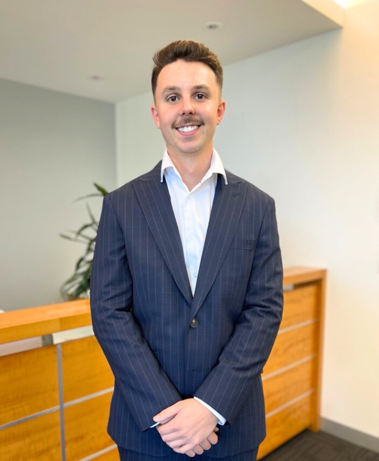 A man in a pinstripe suit stands in an office setting, smiling. He has short dark hair and a mustache, and is positioned in front of a wooden reception desk.