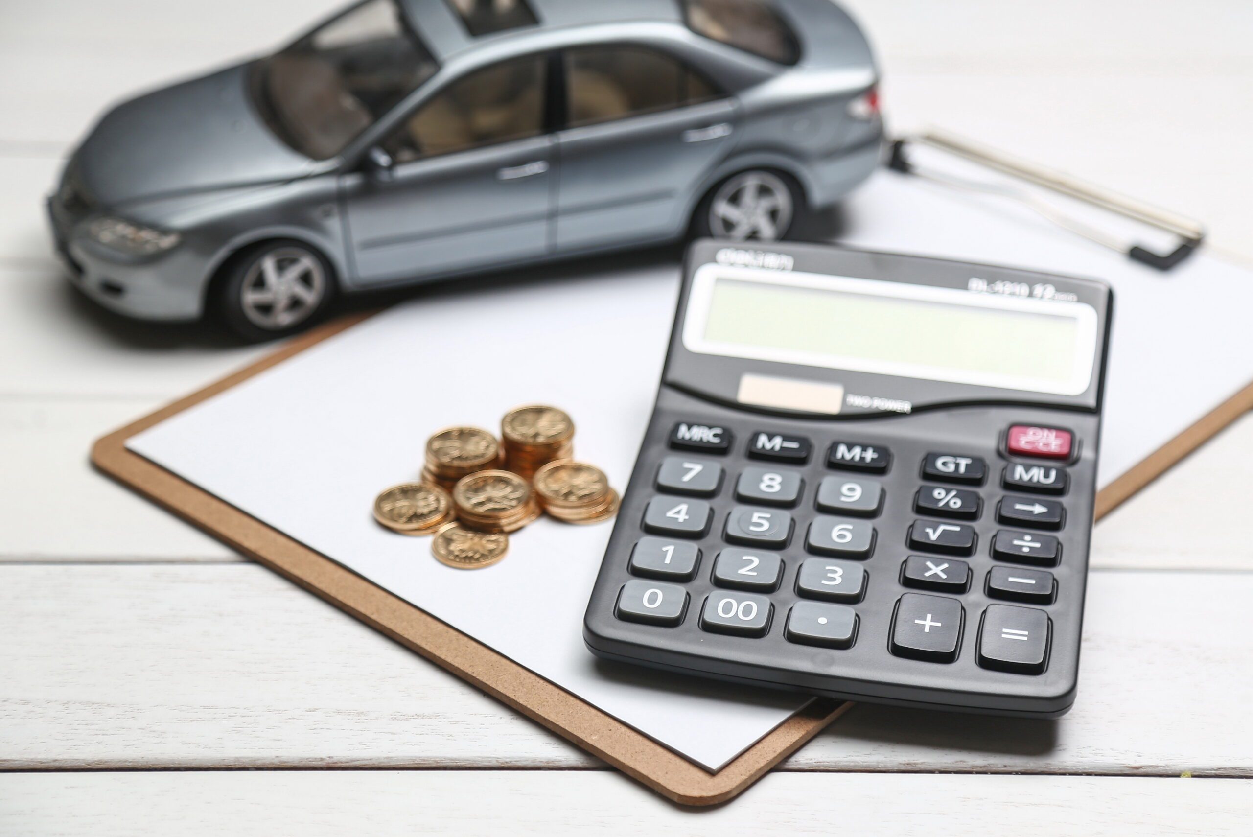 A gray toy car sits on a clipboard with blank paper, next to stacked coins and a black calculator on a white wooden surface. The items subtly hint at financial planning, reminiscent of tools used by business accountants for managing car expenses and budgeting strategies.