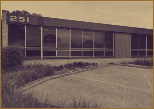 A modern, single-story office building with large tinted windows and the number 251 displayed on the corner, fronted by an empty parking lot and landscaped plants. Image has a sepia tone.