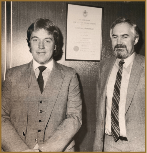 Two men in suits stand and smile in front of a wood-paneled wall with a framed certificate displayed behind them. The photo is black and white, giving a vintage feel.