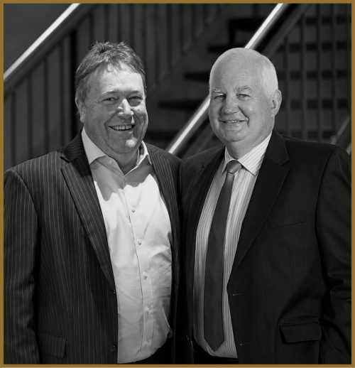 Two smiling men in suits stand side by side in front of a staircase. The image is in black and white, with both men looking at the camera.
