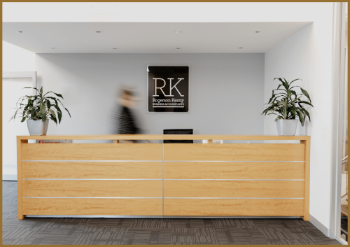 A modern reception desk with a blurred person walking behind it, flanked by two potted plants. A sign reading "RK Rogerson Kenny Business Accountants" is mounted on the wall behind the desk.