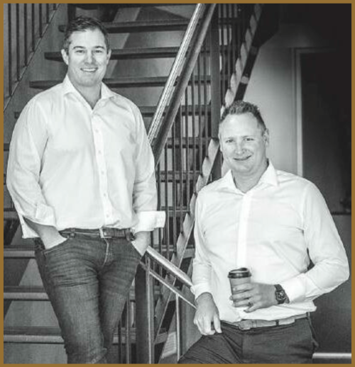 Two men in white shirts and jeans stand by a staircase. One is holding a coffee cup and sitting on the railing, while the other stands with his hands in his pockets. The image is in black and white.