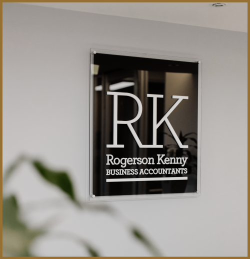 A wall-mounted sign displays the letters "RK" and the words "Rogerson Kenny Business Accountants" in a modern office setting, with a blurred plant in the foreground.