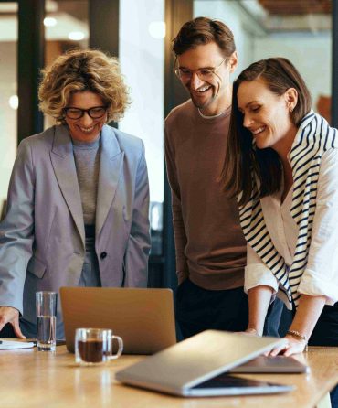 Three people, two women and one man, stand around a table looking at a laptop screen. They are smiling and appear engaged in their discussion. The scene takes place in a modern, well-lit office with glass walls. Coffee mugs, papers, and business accountants’ notes are on the table. Three people, two women and one man, stand around a table looking at a laptop screen. They are smiling and appear engaged in their discussion. The scene takes place in a modern, well-lit office with glass walls. Coffee mugs, papers, and business accountants' notes are on the table.