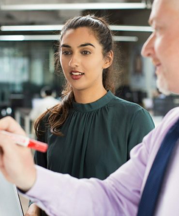 A woman with long hair tied back is attentively looking at a tax accountant who is writing on a glass board with a red marker. The man is slightly out of focus and appears to be explaining something. They are in a modern office setting.
