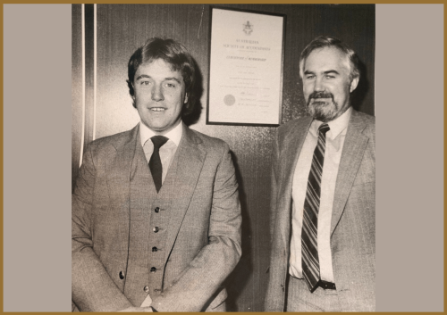 Two men in suits stand side by side, smiling, in front of a plaque or certificate on a wood-paneled wall. The image is in black and white.