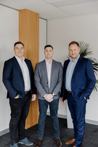Three men in business attire stand together indoors in a modern office setting. They are wearing suits and pose side by side, with a potted plant and wooden panel visible in the background.