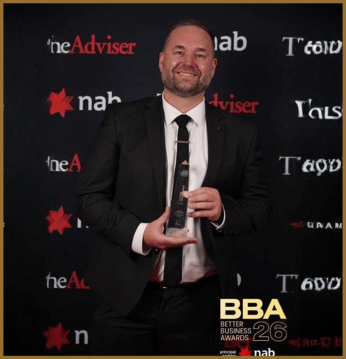 A man in a black suit and tie smiles while holding an award trophy. He stands in front of a backdrop with logos, including "BBA 26 Better Business Awards," "nab," and "theAdviser.
