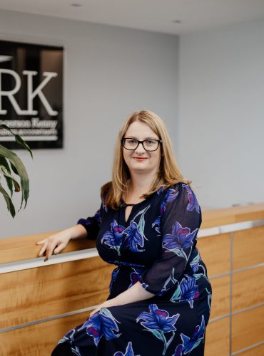 A woman with blonde hair and glasses, wearing a dark blue floral dress, sits on a wooden reception desk in a modern office with a sign behind her that reads: "Align Your SMSF With Your Business Strategy And Maximise Bot.