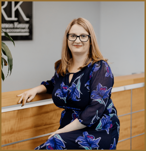 A woman with blonde hair and glasses, wearing a navy blue dress with floral patterns, sits on the edge of a wooden reception desk in an office setting.