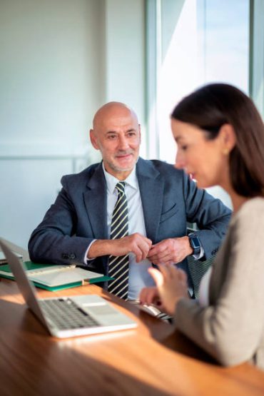 A man in a suit sits at a desk, smiling and looking at a woman working on a laptop. The setting appears to be a modern office with large windows—perfect for Retail Business Accountants Melbourne collaborating on financial solutions.