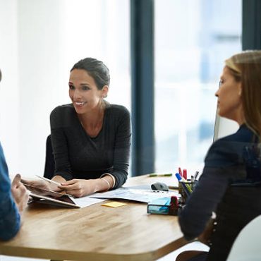 Three people sit around a wooden table in a bright office, engaged in a discussion. A woman in the center is smiling and holding documents, while the other two listen attentively. Papers and office supplies are on the table.