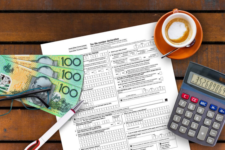 A tax form on a wooden table with three 100 Australian dollar bills, a pair of glasses, a red pen, a calculator, and a cup of coffee in an orange saucer.
