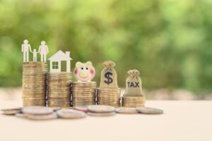 Stacks of coins with paper cutouts of a family and house on top. A pink piggy bank, a money sack with a dollar sign, and another sack labeled "TAX" rest on additional stacks. Blurred green foliage in the background.