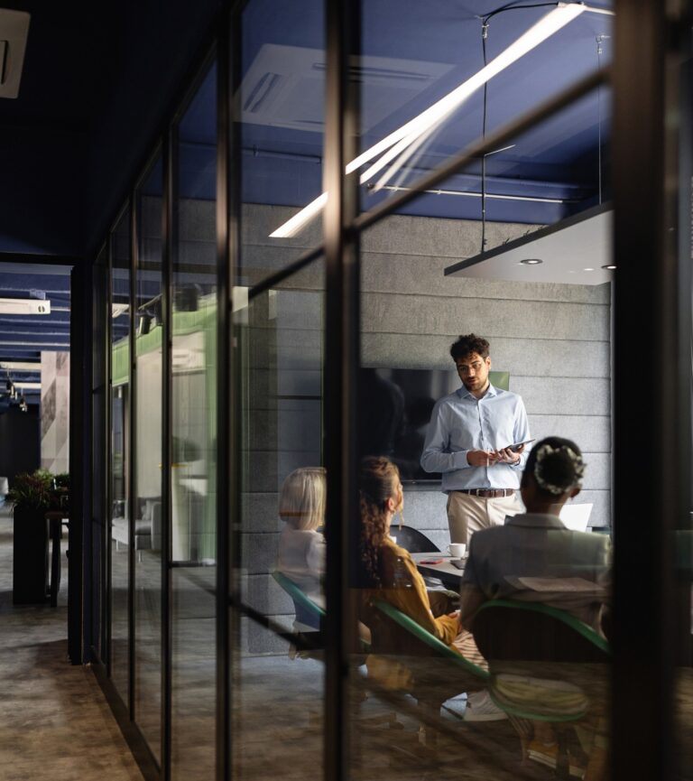 A man stands and presents to three seated colleagues in a modern office meeting room with glass walls and a dark, contemporary interior.