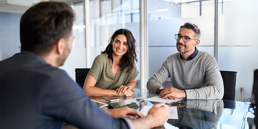 Three people sit at a glass table in a modern office. A woman and a man face a third man, who has his back to the camera. They are engaged in a friendly, professional conversation.