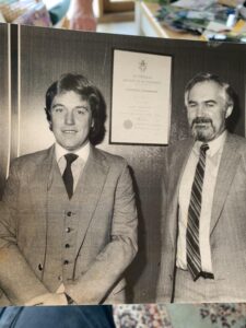 Two men in suits stand side by side in front of a wooden wall with a framed certificate behind them. The photo is black and white and appears to be from the late 20th century.