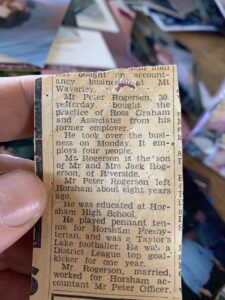 A hand holds an old newspaper clipping featuring an article about Mr. Peter Rogerson, detailing his background, education, and recent business acquisition. Other blurred photos are visible in the background.
