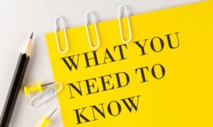A yellow paper with the words "WHAT YOU NEED TO KNOW" in bold black letters, next to two pencils, three paper clips, and three push pins on a white background.