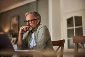 Middle-aged man with glasses and gray hair sits at a table, looking thoughtfully at a laptop screen in a warmly lit room. He rests his hand near his mouth, appearing focused and contemplative.