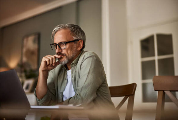 Middle-aged man with glasses and gray hair sits at a table, looking thoughtfully at a laptop screen in a warmly lit room. He rests his hand near his mouth, appearing focused and contemplative.