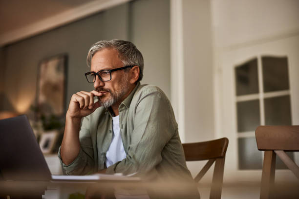 Middle-aged man with glasses and gray hair sits at a table, looking thoughtfully at a laptop screen in a warmly lit room. He rests his hand near his mouth, appearing focused and contemplative.