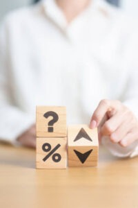 A person in a white shirt stacks wooden blocks with symbols: a question mark, a percent sign, and arrows pointing up and down, suggesting decision-making or financial uncertainty.