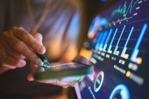 A person uses a stylus on a smartphone, with graphs and data charts displayed on a large digital screen in the background, suggesting financial analysis or stock market monitoring.