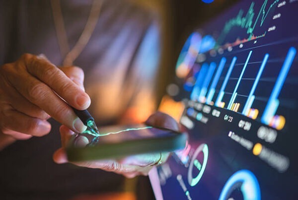 A person uses a stylus on a smartphone, with graphs and data charts displayed on a large digital screen in the background, suggesting financial analysis or stock market monitoring.