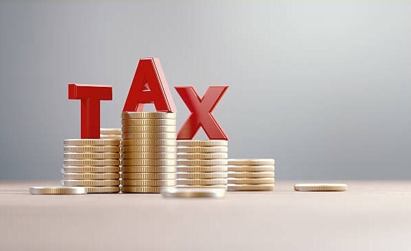 Several stacks of gold coins are arranged on a flat surface, with large red letters spelling "TAX" placed on top of the stacks. The background is plain and light-colored.