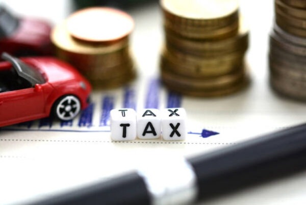 Small blocks spelling "TAX" sit on a document with a pen nearby, stacks of coins, and a red toy car, representing financial and automobile tax concepts.