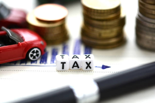 Small blocks spelling "TAX" sit on a document with a pen nearby, stacks of coins, and a red toy car, representing financial and automobile tax concepts.