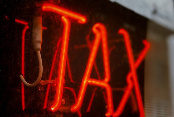 A red neon sign displays the word "TAX" in capital letters. Captured in a slightly dim environment, a reflection dances on the glass while trees and power lines loom in the background, evoking a call for expert Tax Advice amidst urban life.