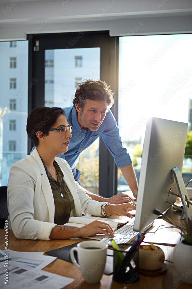 Two people in an office setting at a Melbourne Accounting Firm. A woman with short hair and glasses is seated, working on a computer. A man with curly hair is standing behind her, leaning in to look at the monitor. They are surrounded by office materials and papers, with large windows in the background.