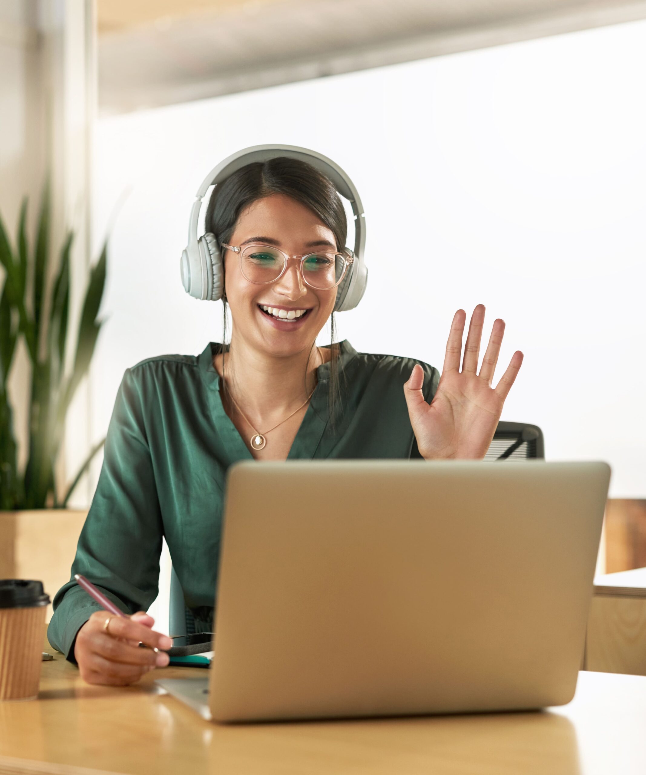 A woman in a green blouse sits at a desk, wearing headphones and glasses, smiling and waving at her laptop screen. She holds a pen, with a notebook and a coffee cup on the desk. A plant is visible in the background as she chats with her business advisor about tax advice.