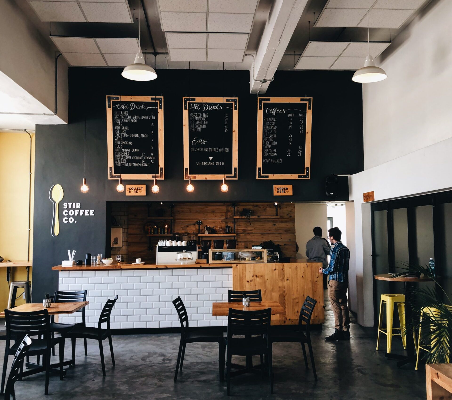 A cozy coffee shop with a modern interior, featuring a white tiled counter, blackboard menus hanging above, and wooden accents. Several tables with black chairs and a yellow bar stool adorn the space. Two people converse about Business Advisors near the counter. "Stir Coffee Co." is proudly displayed on the left wall.