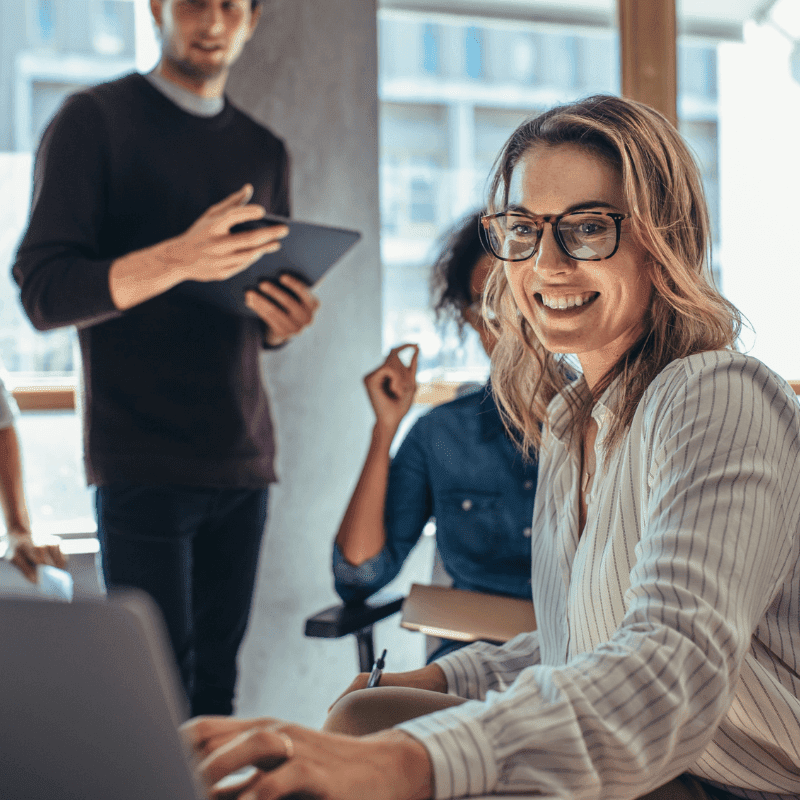A woman wearing glasses and a striped shirt sits at a desk working on a laptop, smiling brightly. In the modern office setting of a Melbourne accounting firm, a man stands in the background holding a tablet while another person is partially visible raising their hand, perhaps seeking tax advice.