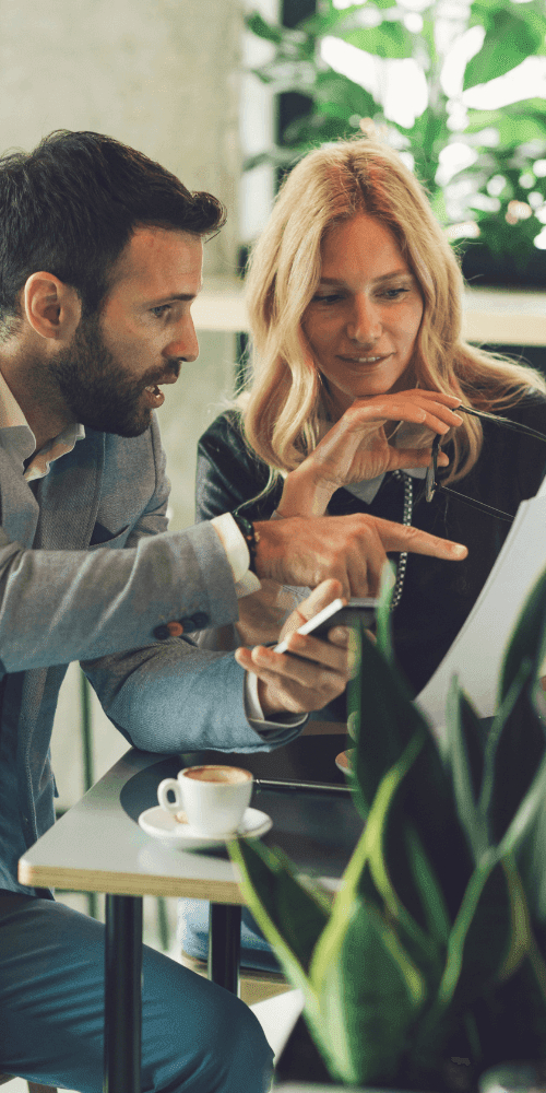 A man and a woman are seated at a small table in a modern setting with green plants in the background. The man, dressed in a light blue jacket, is pointing at a document the woman is holding. They appear to be Business Advisors from a Melbourne Accounting Firm. A cup of coffee and a smartphone are on the table.